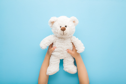Baby boy hands holding smiling white teddy bear on light blue table background. Pastel color. Closeup. Point of view shot. Kids best friend. Top down view.