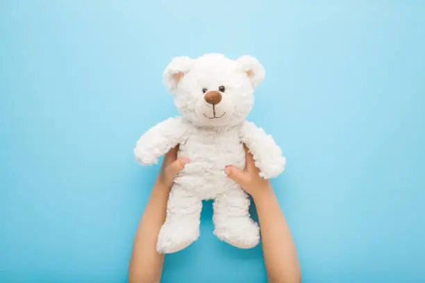 Photo of Baby boy hands holding smiling white teddy bear on light blue table background. Pastel color. Closeup. Point of view shot. Kids best friend. Top down view.