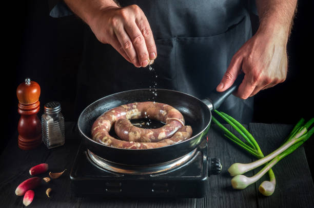 The chef adds salt to pan with raw meat sausage. Preparation for cooking sausages in the kitchen of a restaurant or cafe on table with vegetables and spices