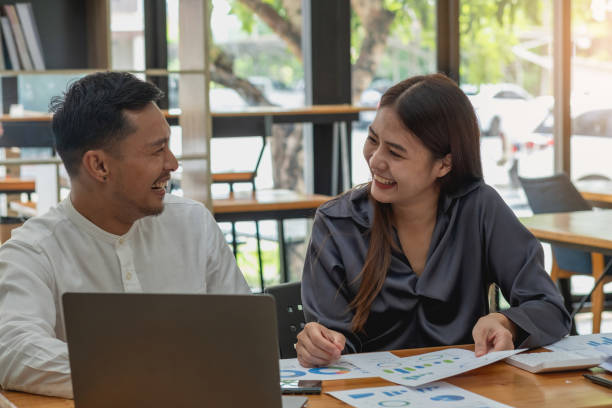 Two male and female marketers use company documents and use laptop computers to record data to analyze company profits.