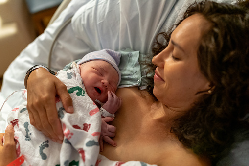 A beautiful multiracial woman lying in a hospital bed smiles affectionately at her newborn baby who is sleeping peacefully in her comforting embrace as they enjoy bonding with skin to skin contact.