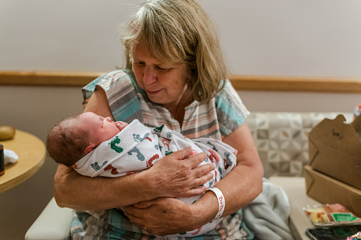 A mature adult Caucasian woman sits in the labor and delivery room at the hospital and affectionately cradles her adorable Eurasian newborn grandchild who is swaddled in a receiving blanket and sleeping peacefully in her comforting embrace.