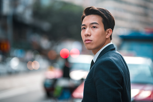 Serious young Asian male lawyer standing outdoors in the city. He is wearing a formal suit and a tie. He is on his way to meet a client.