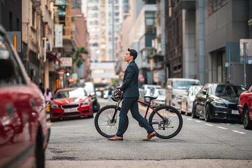 Attractive young Chinese male lawyer and entrepreneur walking in the city. He is carrying a bicycle while crossing the street from right to left. He is wearing a formal suit.