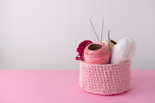 Chunky yarn basket with yarn balls, knitting needles and a crochet hook on pastel pink table, copy space