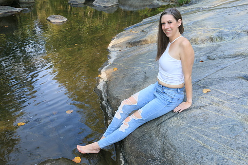 A Caucasian woman sitting on a rock enjoying the peace and quiet. She is wearing a white short sleeveless top, torn jeans and is barefoot.
