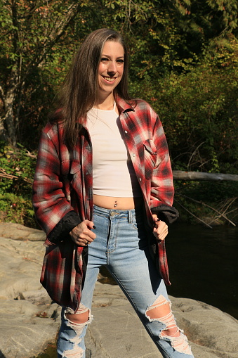 A Caucasian Canadian woman on a rock beside a river in Autumn. She is wearing a short white top, torn jeans and an open plaid shirt.