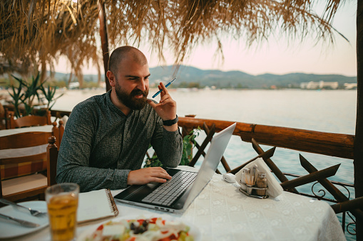 Young bearded man using a laptop and studying in a seaside restaurant during a summer vacation in Greece