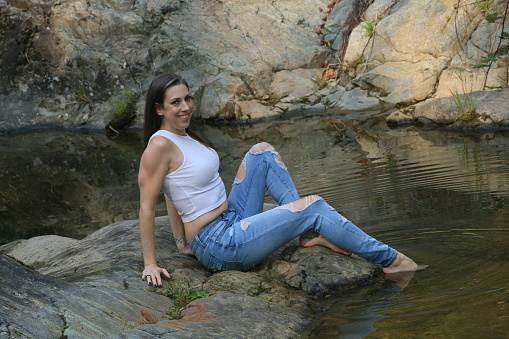 A Caucasian woman with her toe in a river in Autumn. She is wearing a white sleeveless top and torn jeans with barefeet.