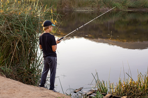 Teen boy with fishing rod standing on the lake shore in the evening light, looking at the water