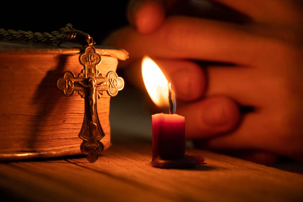 A woman holds a cross near a candle and a bible in the dark, a woman prays, praying, faith and religion stock photo