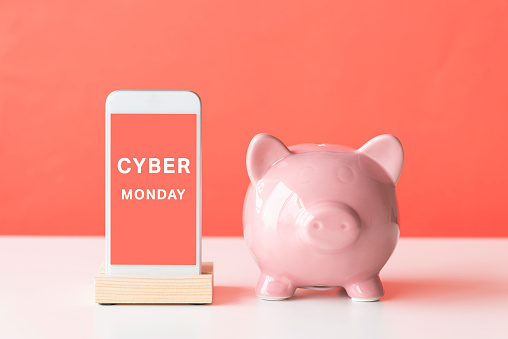 Front view of a smart phone and pink piggy bank on white table in front of a red background. Cyber Monday text on device screen.