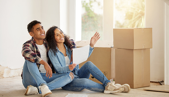 Couple happy on the floor of their new property or house in the living room with box in the background. Real estate owner smile inside the lounge interior thinking of idea for renovation at home