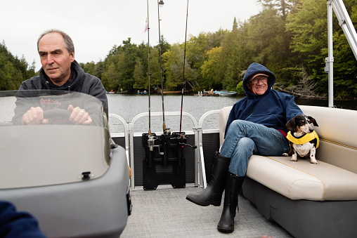 Two senior men enjoying a pontoon boat tour on a lake in autumn. They are family, one is in his fifties and other in his sixties. They are wearing warm clothes. Dachshund dog wearing a life vest near the oldest man. Horizontal outdoors waist up shot with copy space.