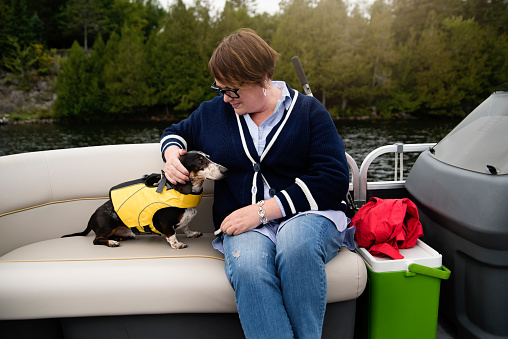 Senior woman and a dog enjoying a pontoon boat tour on a lake in autumn. She is wearing warm clothes. Dachshund dog wearing a life vest. Horizontal outdoors full length shot with copy space.