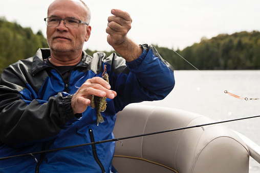 Senior man fishing a pontoon boat tour on a lake in autumn. Man is wearing warm clothes. Fish is too small and will go back in water. Horizontal outdoors waist up shot with copy space.