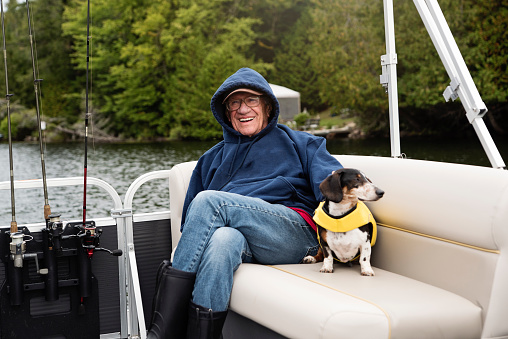 Senior man and a dog enjoying a pontoon boat tour on a lake in autumn. Man is wearing warm clothes. Dachshund dog wearing a life vest. Horizontal outdoors full length shot with copy space.