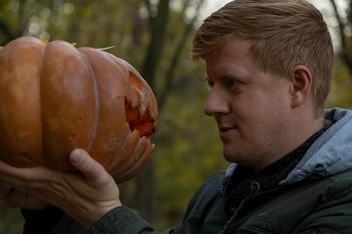 Close-up view of caucasian man holding orange carved Halloween pumpkin and looking into Jack's eyes in dark autumn forest. Face to face. Holiday decoration theme.