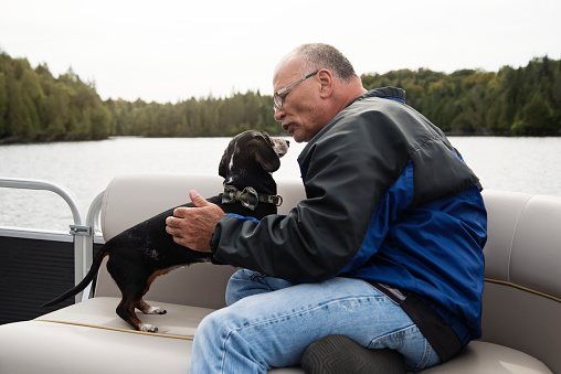 Senior man and a dog enjoying a pontoon boat tour on a lake in autumn. He is wearing warm clothes. Dachshund dog is a rescued dog and is half-blind. Horizontal outdoors full length shot with copy space.