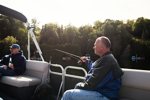 Senior father and mature son enjoying fishing on a pontoon boat tour on a lake in autumn. They are family, senior dad is in his eighty, son in his fifties. They are wearing warm clothes. Horizontal outdoors waist up shot with copy space.