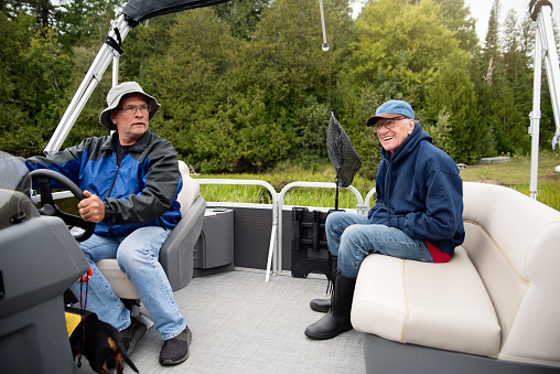 Senior father and mature son enjoying a pontoon boat tour on a lake in autumn. They are family, senior dad is in his eighty, son in his fifties. They are wearing warm clothes. Horizontal outdoors full length shot with copy space.
