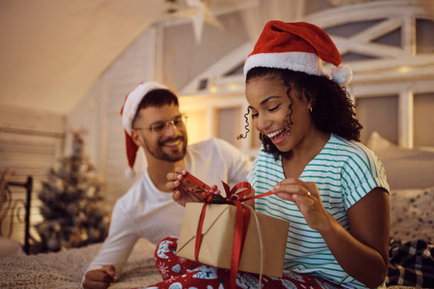 Cheerful black woman opening present she receiving from her boyfriend on Christmas morning. Young African American woman having fun while unwrapping Christmas present from her boyfriend at home. unwrapping stock pictures, royalty-free photos & images