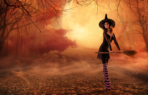 ballerina on pointe shoes in black witch costume with broom is dancing on an old road against the backdrop of huge red moon.