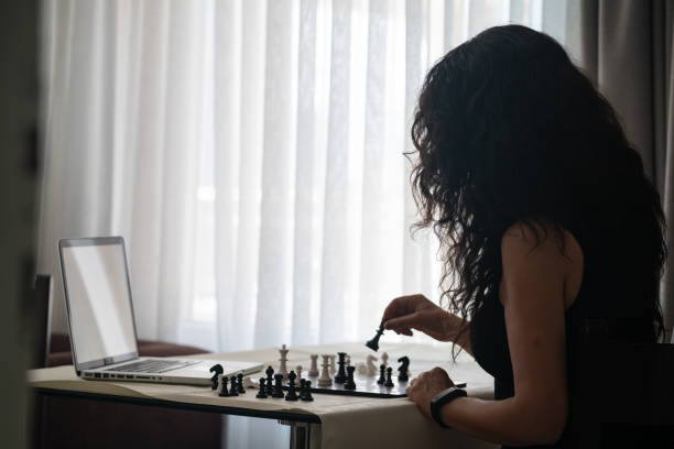 Photo of adult woman learning to play chess online Over the shoulder view of adult woman with long curly hair e-learning playing chess. She is sitting on table next to window. Shot under daylight with a full frame mirrorless camera. computer chess stock pictures, royalty-free photos & images