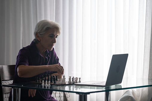 Photo of senior adult woman with white hair sitting on table and using laptop computer for learning playing chess. She is sitting next to window. Shot under daylight with a full frame mirrorless camera.