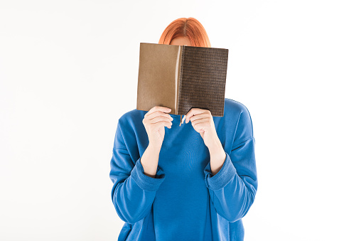 A young and attractive Caucasian redhead girl in a blue jumper covers her face with an interesting book and reads isolated on a white studio background.