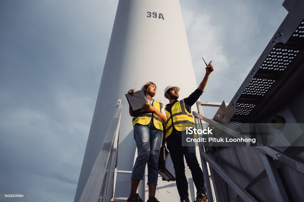 Young man and woman maintenance engineer team working and holding the report in wind turbine farm. Generator station, renewable energy Engineer Stock Photo