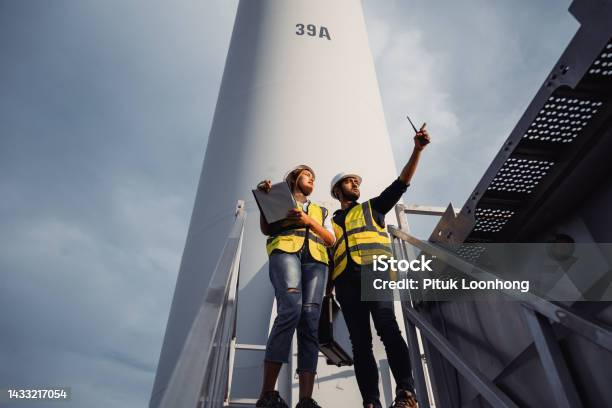 Young Man And Woman Maintenance Engineer Team Working And Holding The Report In Wind Turbine Farm Generator Station Renewable Energy Stock Photo - Download Image Now