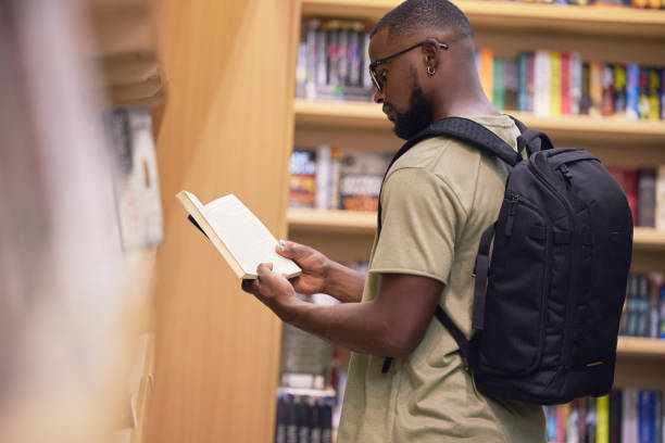 istock Education student with book in a library at university, college or school reading or doing research. Scholarship, learning and study knowledge and black man with print books on shelf and backpack 1433217372