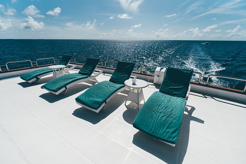 A row of four deckchairs with teal cushions on them and two small coffee tables in between on the upper deck of a luxurious diving safari boat or a recreational vessel lit by bright Maldivian sun
