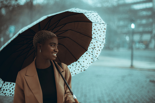 A portrait of a very beautiful youthful smiling black female looking aside while hiding from the heavy rain under her huge spotty umbrella while standing on the misty bluish street on the pavement