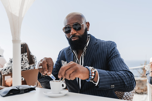 A portrait of a dapper mature hairless black guy with a neat beard sitting in a street coastal restaurant and pouring sugar into his espresso coffee cup; an elegant African guy in an outdoor cafe