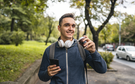 One young adult caucasian man walking in the city or town near park with cup of coffee and mobile phone in autumn or spring day happy male tourist standing alone real people copy space