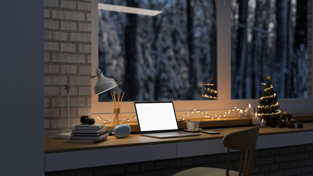 Working space with laptop mockup, Christmas tree, Christmas lights on table against the window Comfortable home working space with laptop white screen mockup, table lamp Christmas tree, Christmas lights, candles and stuff on table against the window with snow view. 3d render, 3d illustration office window night stock pictures, royalty-free photos & images