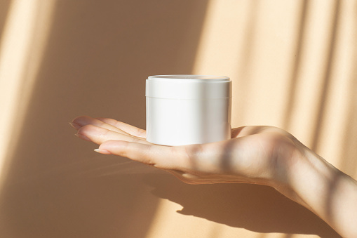 Mockup of white jar for packaging cosmetics on woman's hand. Unmarked container with moisturizer close-up on beige background, in rays of sunlight. Concept of skin care, rejuvenation.