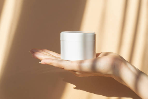 mockup of white jar for packaging cosmetics on woman's hand. unmarked container with moisturizer close-up on beige background, in rays of sunlight. concept of skin care, rejuvenation - moisturizer cosmetics merchandise human hand imagens e fotografias de stock