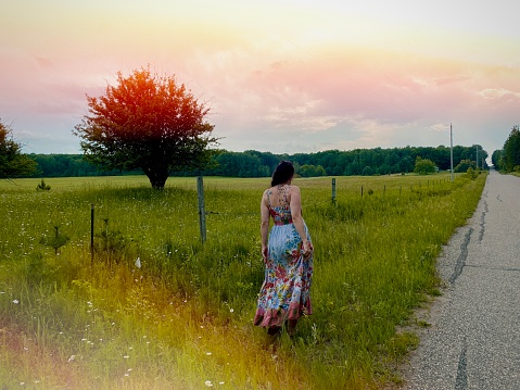 Behind view of beautiful women in a flowered purple sundress, walking away from the camera on the side of a beautiful country road. It is summer and the evening light is streaming surrounding the green trees and fields. Taken in Northern Wisconsin, USA.