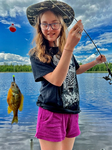 Cute teen girl in large fishing hat, holding her fishing pole out with a caught fish, a small sunfish or pumpkinseed perch, she is smiling and having fun, standing on a dock on a small lake, Cotas lake in Pembine, Wisconsin. Beautiful blue skies and lake covering the background.