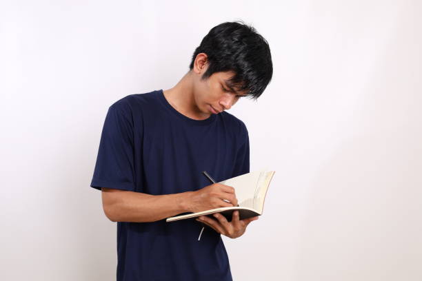 istock Young asian man standing while writing on a book. Isolated on white background with copyspace 1433266950
