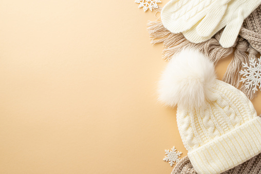 Winter concept. Top view photo of white knitted mittens bobble hat cozy scarf and snowflakes on isolated beige background with copyspace