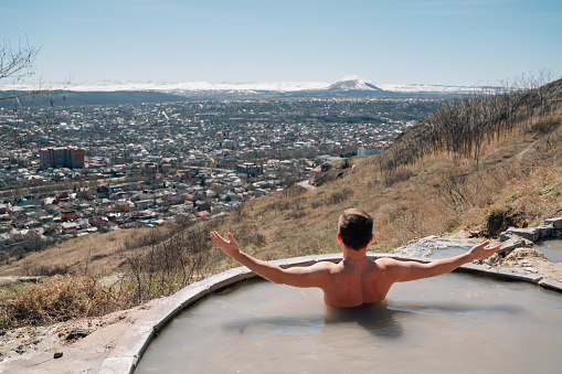 view from the back. a man bathes in a hydrogen sulfide thermal spring on the mountainside against the backdrop of the city. therapeutic and relaxing baths in nature at any time of the year.