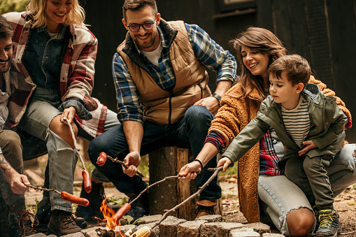 Image of a firepit and happy smiling friends and family of three, father, wife, and son, burning the sausage by the fire and enjoying time together in the background