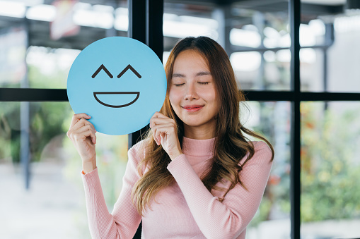Asian woman holding blue happy smile face on paper cut, user giving good feedback rating, think positive , customer review, assessment, of mental health day, Compliment Day, satisfaction concept.