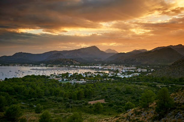 Elevated view from nearby hills of sailboats in Port de Pollença’s harbor and marina, light reflecting on water from clouds over Serra de Tramuntana range.