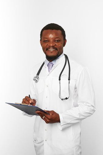 Smiling black bearded doctor man in white robe with stethoscope filling medical records on clipboard, isolated on white background. Happy adult black african american physician therapist portrait