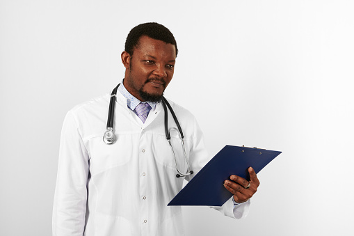 Black bearded doctor man in white robe with stethoscope holds medical records on clipboard, isolated on white background. Serious adult black african american physician therapist portrait